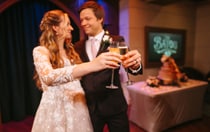 A bride and groom smiling and toasting champagne glasses in front of a tiered wedding cake at The Bayou bar on a Disney Cruise Line ship