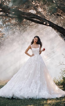 A woman posing in a forest wearing a fairy tale wedding gown with floral details holding an apple