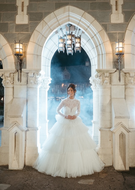 A bride standing in an archway while wearing a wedding gown featuring a tiered skirt and a beaded illusion neckline and sleeves