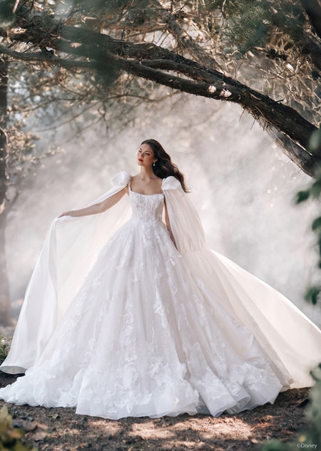 A woman posing in a forest wearing a fairy tale wedding gown with floral details and a flowing cape with puffed sleeves 