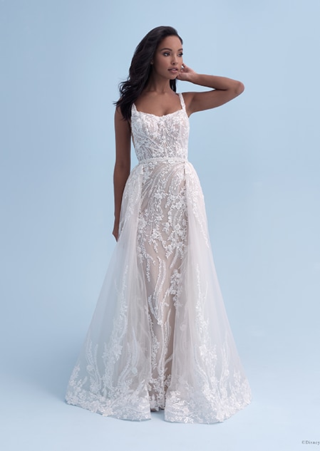 A bride in a form fitting wedding dress with a square neckline decorated with crystal flowers, with additional layers added onto the sides of the skirt A bride in a form fitting wedding dress with a square neckline decorated with crystal flowers, with additional layers added onto the sides of the skirt