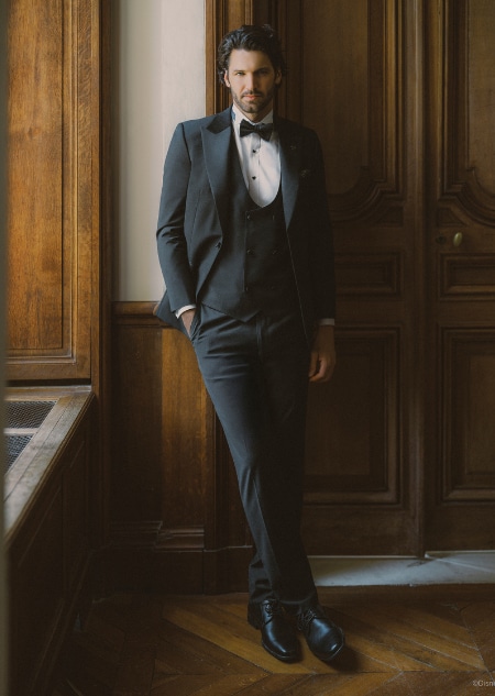 A man in a black tuxedo and bow tie smiling while sitting near floral arrangements  
