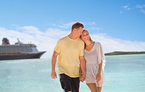 A couple holding hands while walking on a beach with a Disney cruise ship behind them at Castaway Cay