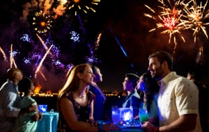 A couple enjoying beverages at an outdoor cocktail table as fireworks burst above them