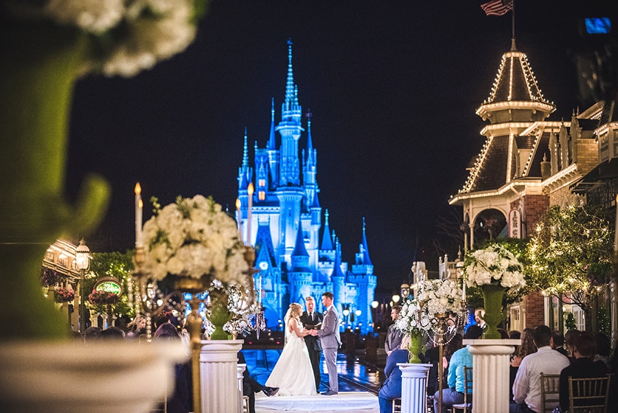 A couple and an officiant stand in front of Cinderella Castle as they get married on Main Street, USA which is decorated with columns of floral bouquets and has a group of seated Guests
