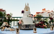 A two tiered wedding cake with a Cinderella Castle cake topper on a table adorned with flowers and silver tea lights 