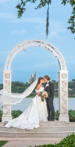 A wedding couple kissing under a trellis near Seven Seas Lagoon and Cinderella Castle in the distance A wedding couple kissing under a trellis near Seven Seas Lagoon and Cinderella Castle in the distance