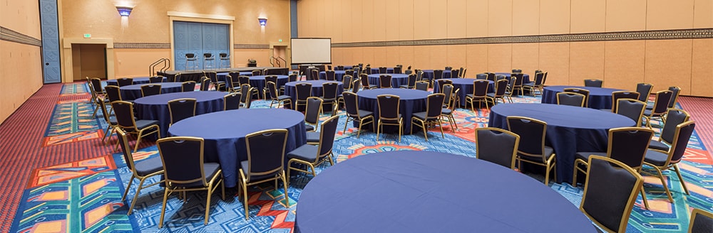 Several circular tables surrounded by chairs arranged in a room with a stage and a screen