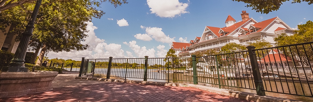A shady brick path with a cast iron guardrail and old fashioned streetlamps looks out to the Seven Seas Lagoon and Disney's Grand Floridian Resort & Spa