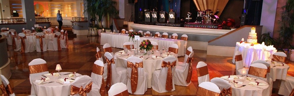 A ballroom with a stage for a live band, tables topped with floral bouquets and dinnerware, chairs decorated with ribbon backs and a wedding cake