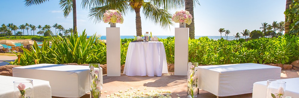 In a beachfront garden at Aulani Resort, banquettes and glass vases are arranged in front of an altar that is flanked by pillars topped with rose bouquets