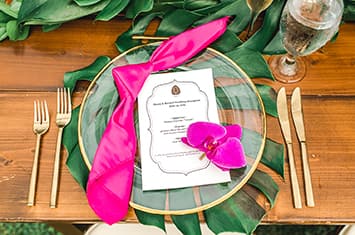 A reception table setting with an itinerary, an orchid, a ribbon, cutlery, a wine glass and clear plate atop a large leaf