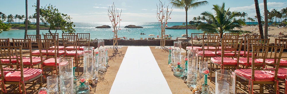 An outdoor wedding reception decorated with 2 trees at the front, aisles of chairs on either side, glass vases along the carpet path and a view of the ocean and palm trees
