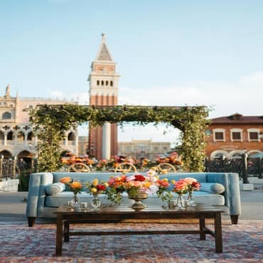 A sofa in front of a wedding arch in a plaza and a coffee table with 5 floral arrangements in vases