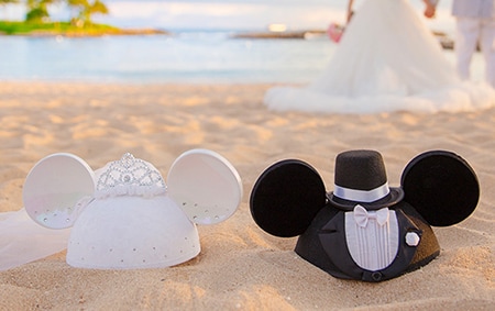 A Minnie ear hat decorated with a crown and veil next to a Mickey ear hat decorated with a top hat and tuxedo