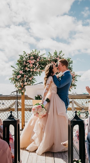 In front of a floral arrangement on the deck at The Wedding Pointe at Castaway Cay, a couple in a tux and bridal gown embrace In front of a floral arrangement on the deck at The Wedding Pointe at Castaway Cay, a couple in a tux and bridal gown embrace