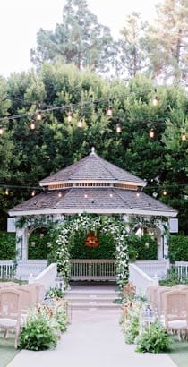Amid strings of decorative lights, chairs are arranged on a lawn to face a gazebo adorned with flowers Amid strings of decorative lights, chairs are arranged on a lawn to face a gazebo adorned with flowers
