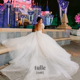 A bride in a wedding dress with a large tulle train, standing in front of Sleeping Beauty Castle