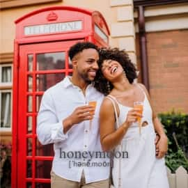 A newly wed couple smiling and embracing while holding drinks in front of a British telephone booth on their honeymoon