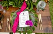 A wedding dinner table setting with an itinerary card, an orchid blossom, a tied ribbon, gold utensils, a wine glass and a clear glass plate sitting on a large leaf