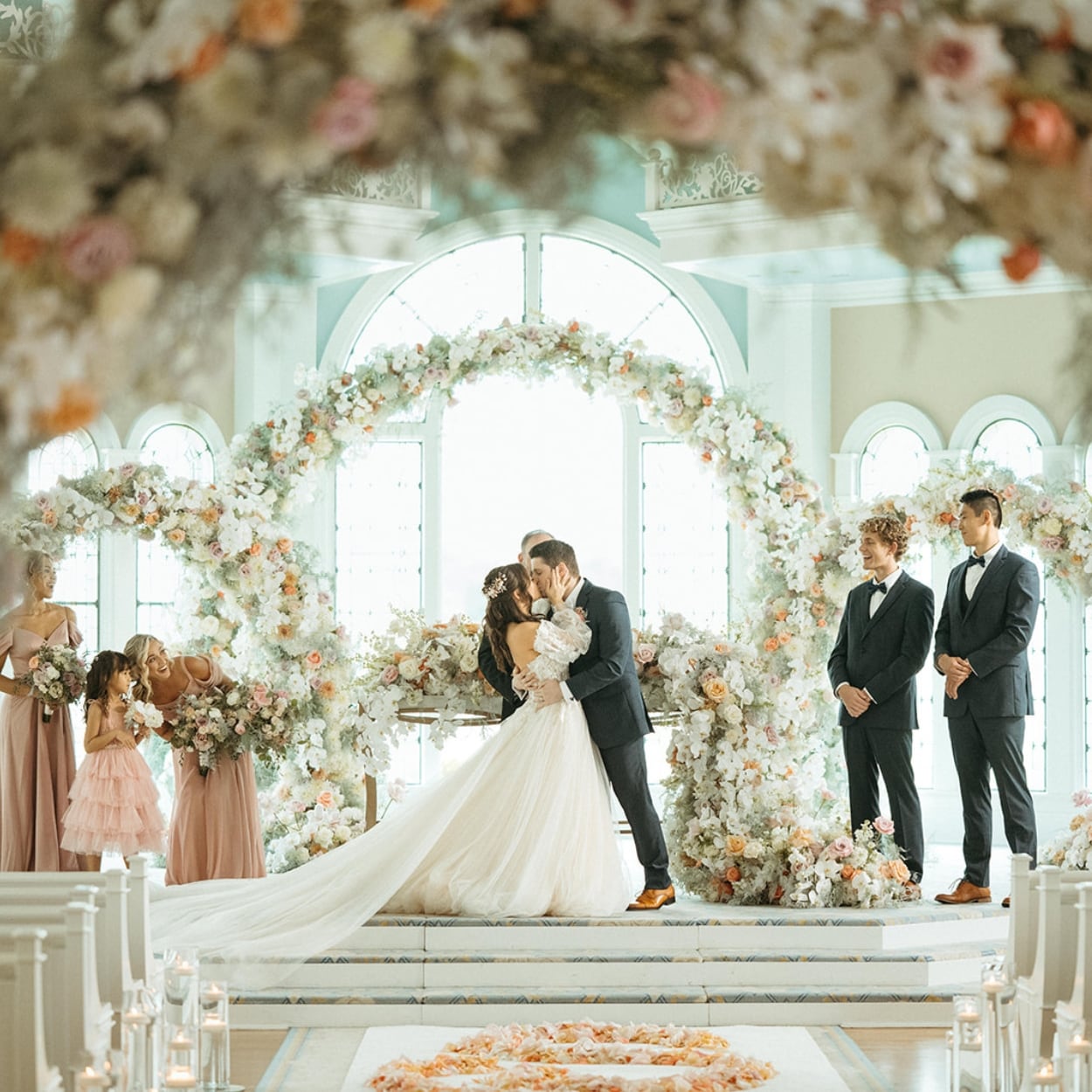 A wedding couple kissing under a floral arch with the wedding party watching