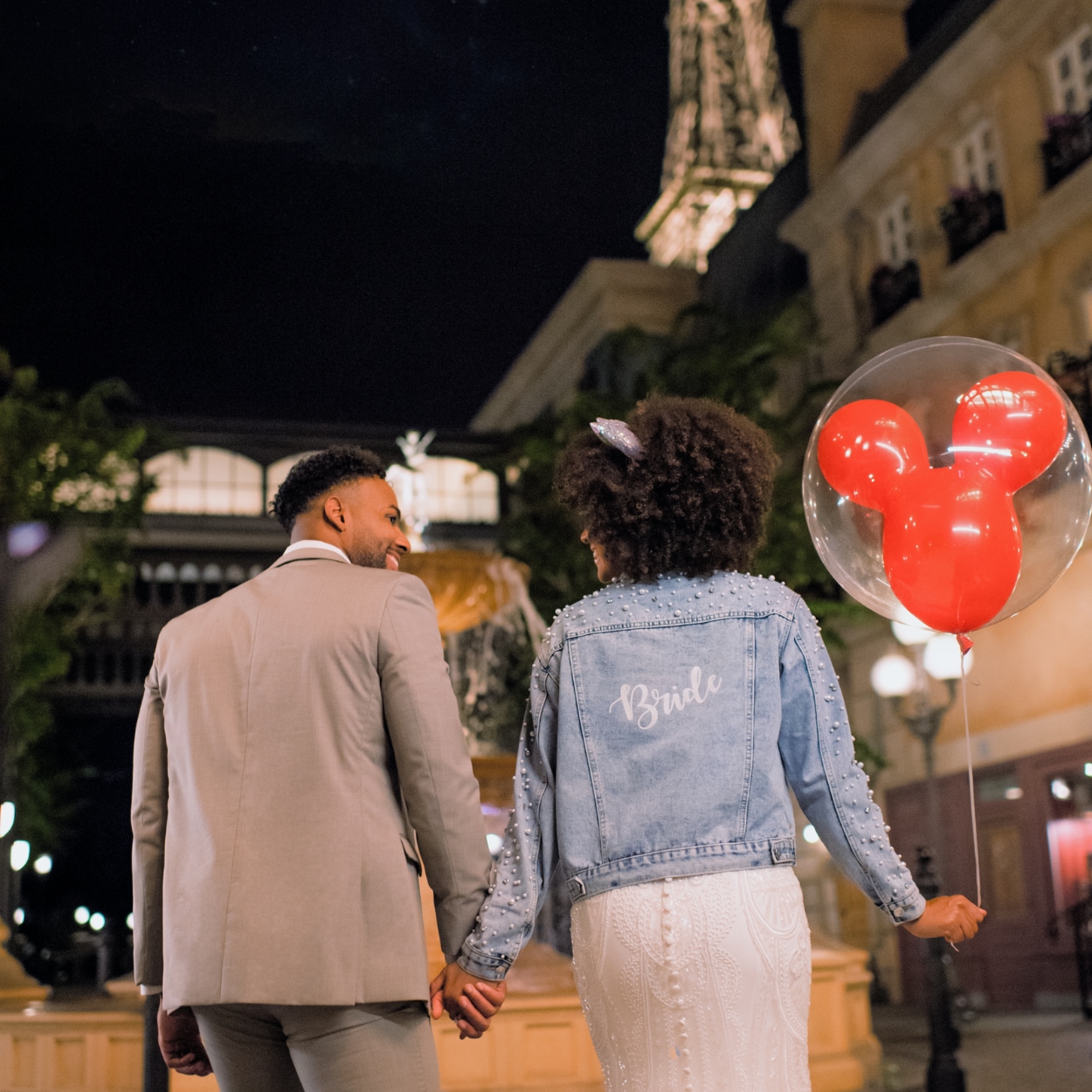 A woman wearing a wedding dress and a jacket that says ‘Bride’, holding hands with a man while carrying a Mickey Mouse balloon