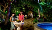 Two women with drinks talk while standing at an outdoor cocktail table near a candlelit stone path
