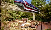 Cast members set up buffet stations for an outdoor event as a monorail glides by overhead
