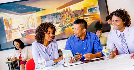 Group of Guests seated at a meeting table with glasses of water, a tablet and mural in the background
