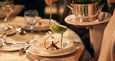 A server pouring soup into a bowl at a formal dining table set with glassware and utensils