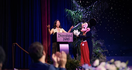 A woman speaking at a podium and Mickey Mouse clapping on stage with the words ‘Disneyland Hotel’
