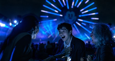 Guests laughing at an outdoor evening event with Mickey’s Fun Wheel illuminated in the background