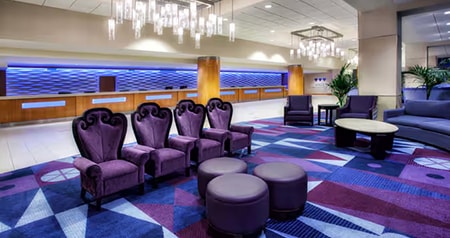 A hotel lobby with ornate armchairs, round ottomans, a geometric patterned carpet and chandeliers
