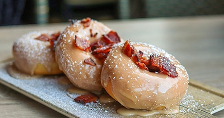 Glazed donuts topped with powdered sugar and bacon pieces served on a wooden board