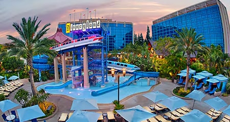 The pool area at the Disneyland Hotel with monorail themed water slides, lounge chairs, umbrellas and hotel towers at sunset
