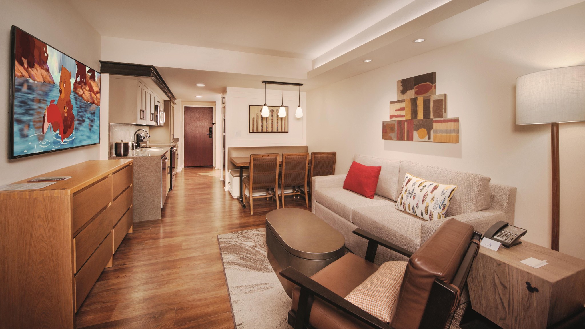 Living area with sleeper sofa, armchair, coffee table with storage, dining table and chairs, hanging accent lights, artwork, dresser, flat panel TV and kitchenette.