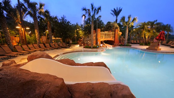 A swimming pool lit up at night with lounge chairs and palm trees surrounding it