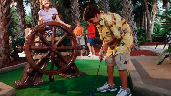 A girl leans on a wooden wheel watching her son putt on a miniature golf course
