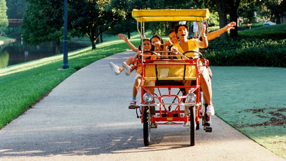 A family of 4 delight in riding a surrey bike along a path flanked by lawn and foliage