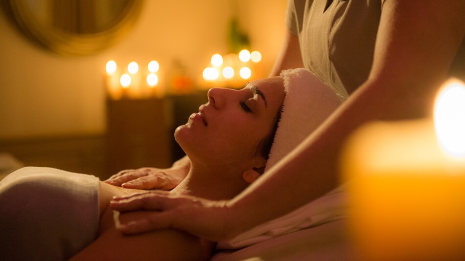 A woman with her hair in a towel lies on her back while her shoulders are massaged