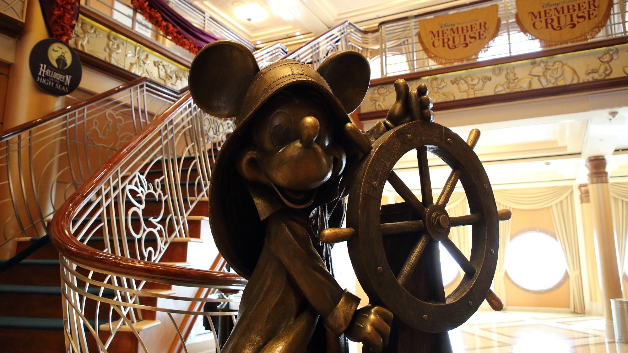 A statue of Mickey Mouse holding a ship's helm, located near a staircase leading up to a balcony that's decorated with banners that read 'Member Cruise'