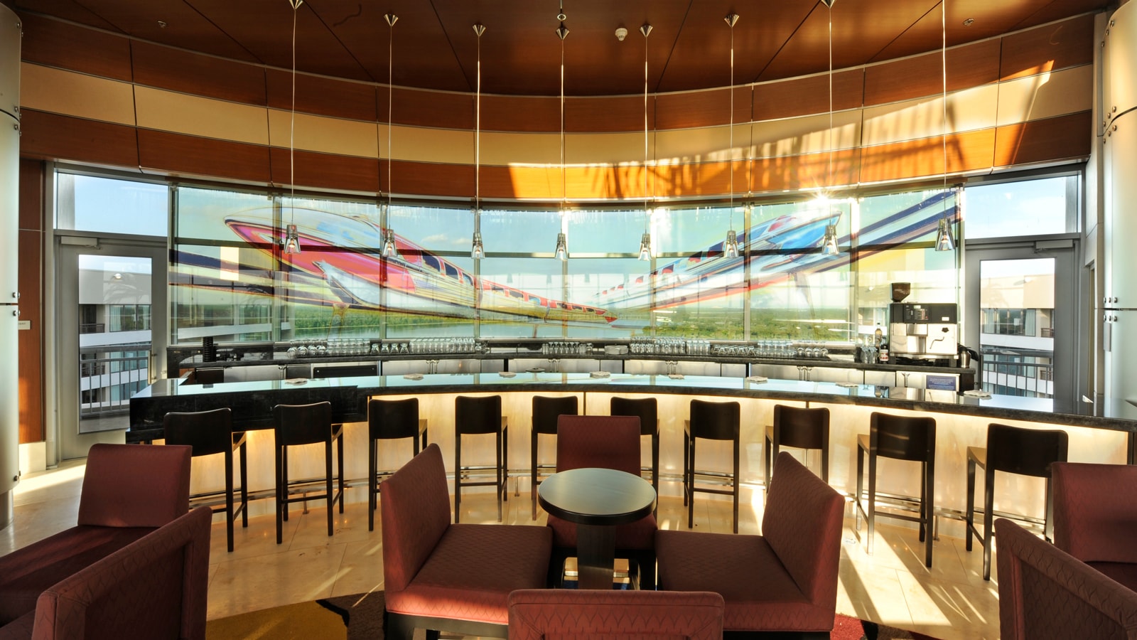 Small tables and chairs arranged in front of a concave bar overlooking glass windows with a monorail view