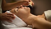 A woman lying face-up on a spa table while a spa therapist applies a brush to her face
