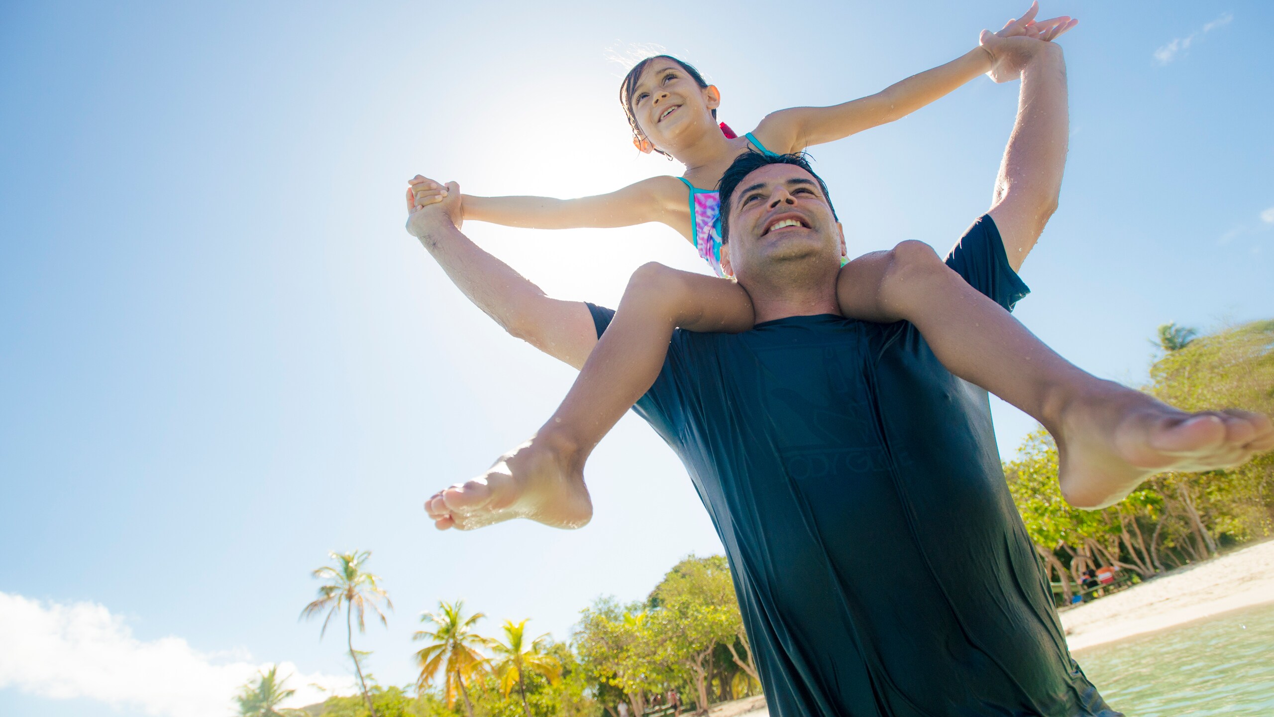 A girl sitting on her dad's shoulders in a tropical beach location