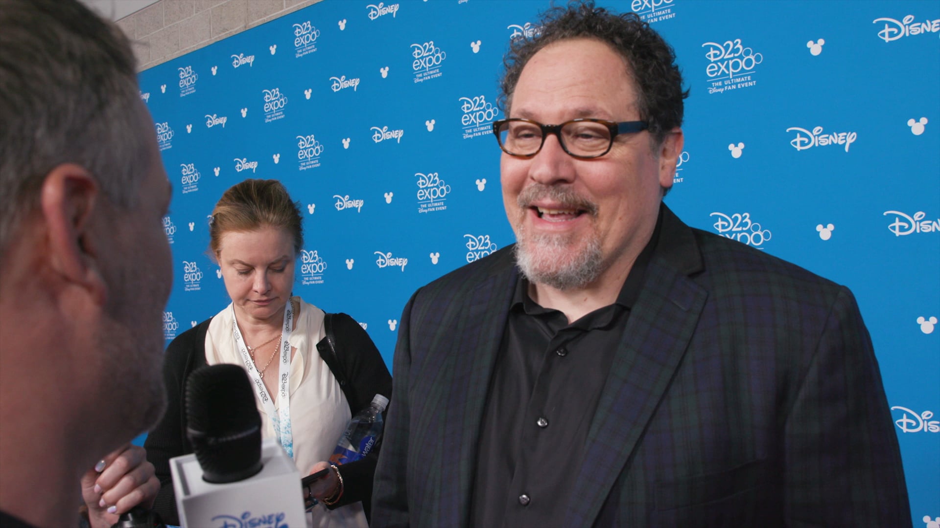 A man interviewing filmmaker Jon Favreau in front of a backdrop with logos that read ‘D23 Expo’ and ‘Disney’