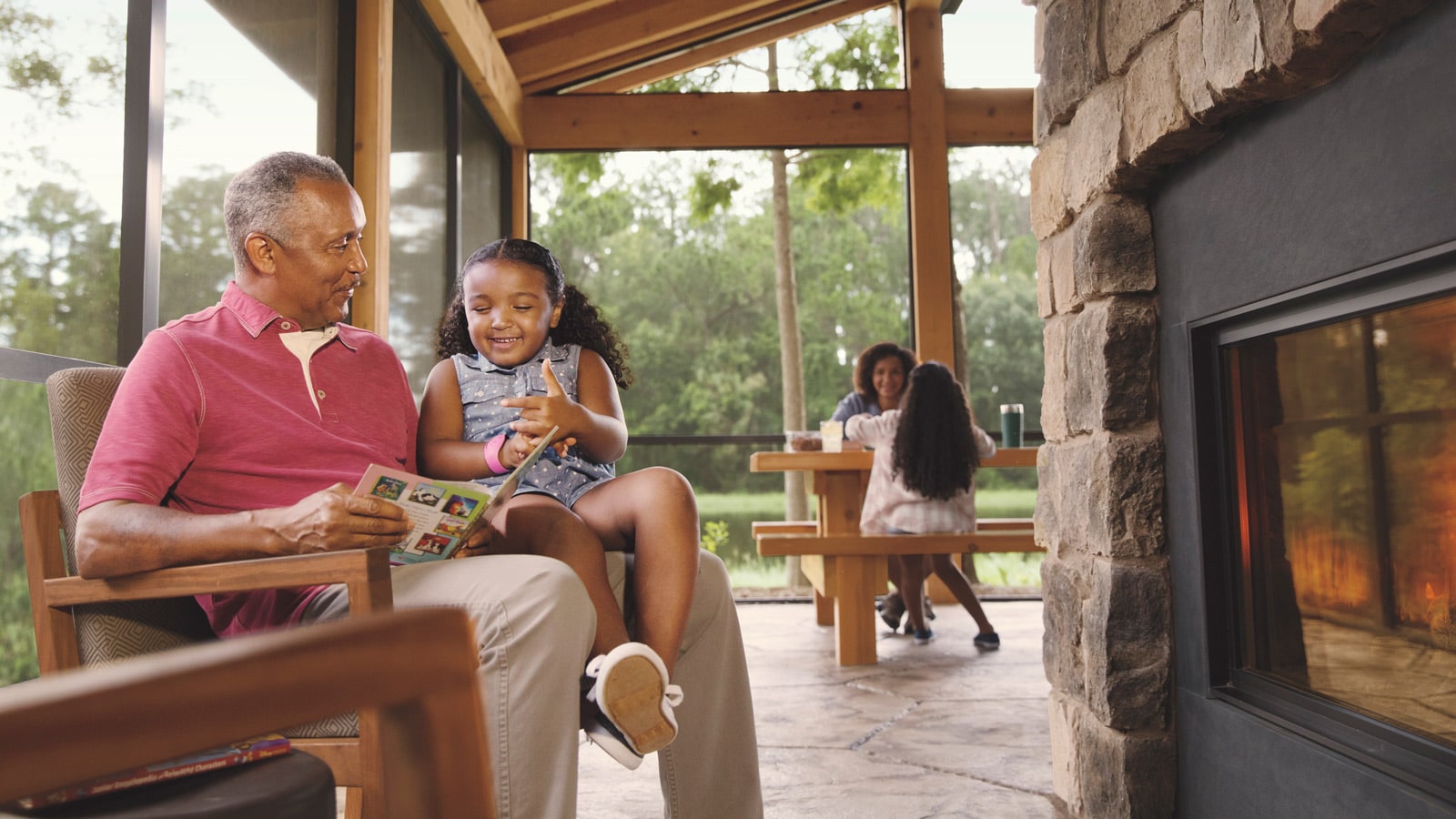 A man reads to a young girl in front of a fireplace and a woman and girl sit at a picnic table on a porch
