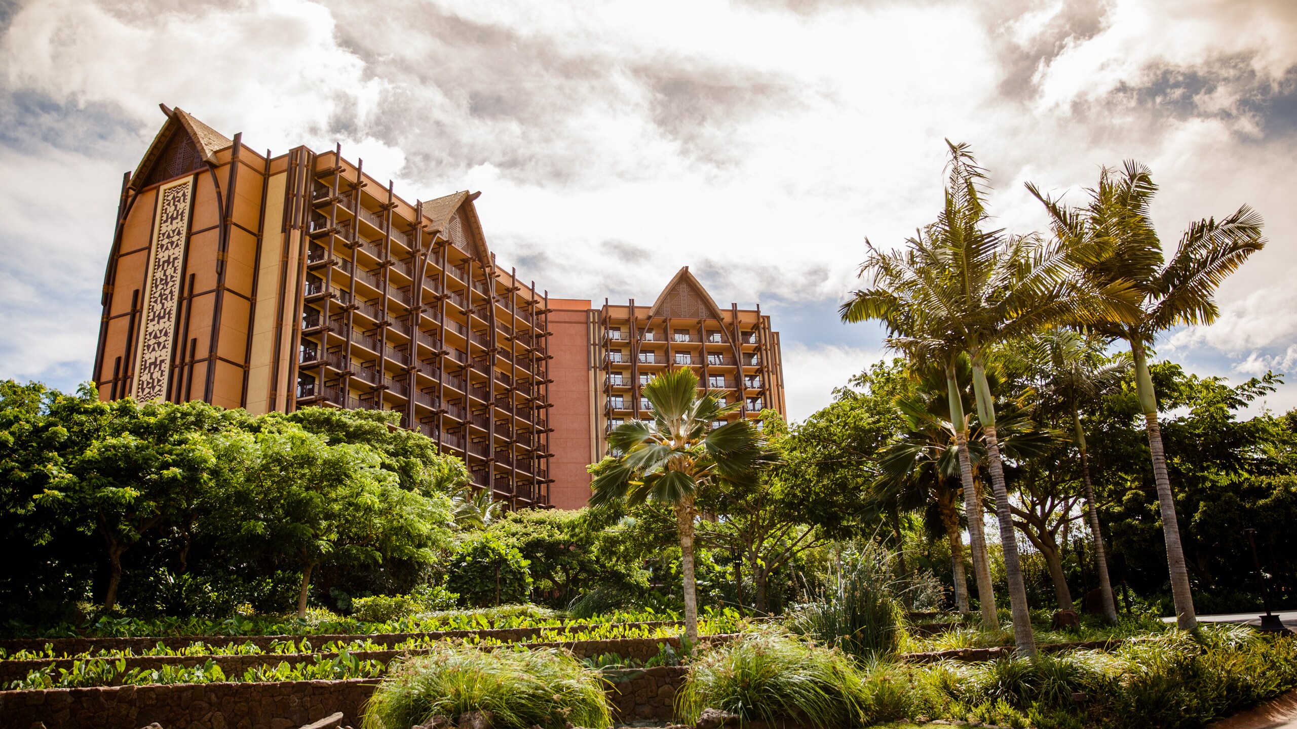 Clouds cover the sky over a tall Hawaiian style resort building surrounded by lush foliage and palm trees