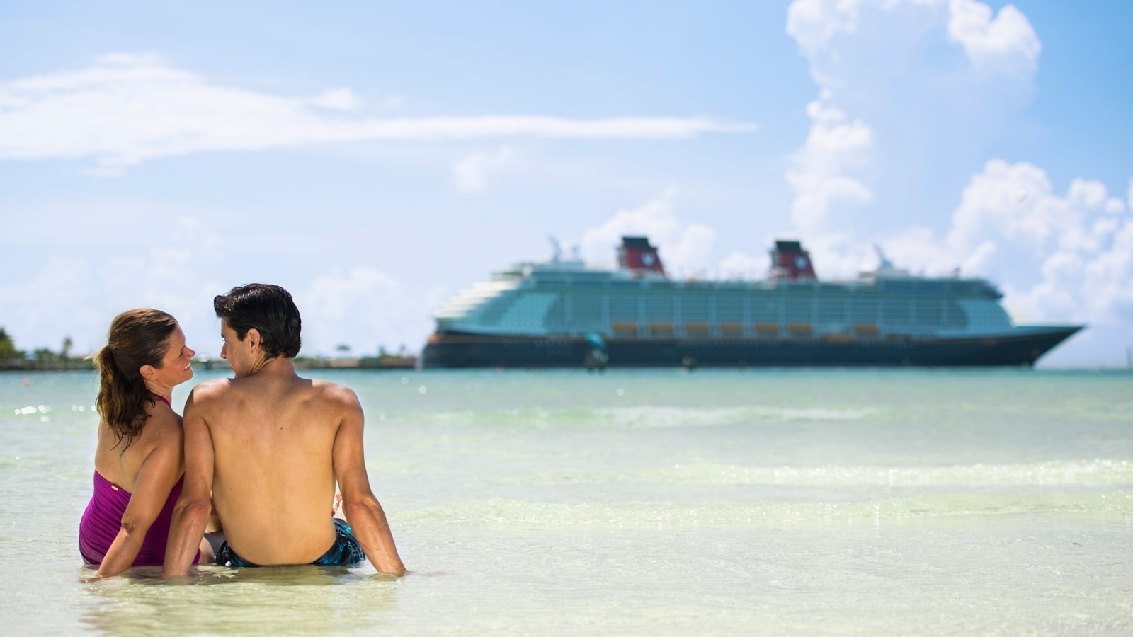 A man and woman lounging in shallow water near a Disney Cruise Line ship