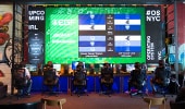 A row of competitors sit at desks in front of individual monitors and a large scoreboard