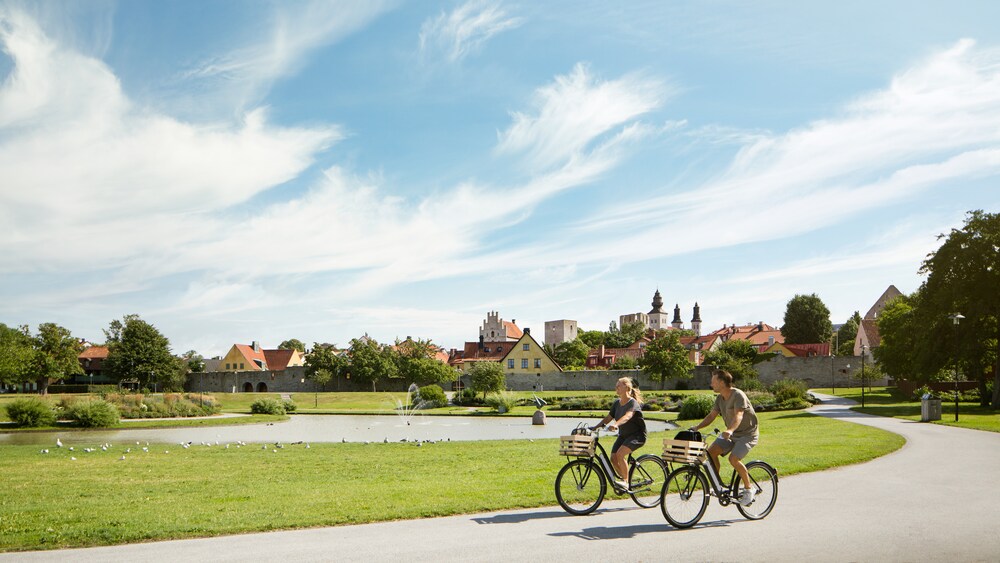 A male and female ride bikes past a small pond with a water fountain